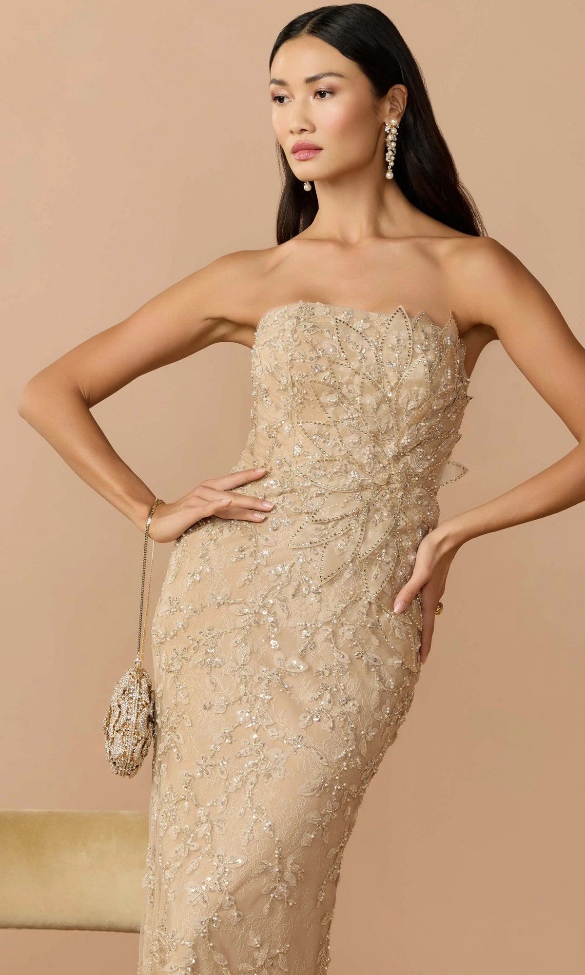 A woman poses in the Mac Duggal 21200 long formal dress, a gold strapless sheath gown with floral embellished lace details. She holds a matching clutch and wears drop earrings, standing against a beige background for an elegant look.